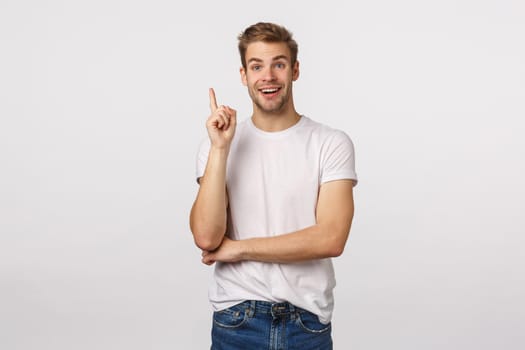 Hear me out, excellent idea striked me. Attractive happy smiling blond guy in white t-shirt, raise index finger eureka gesture and grinning, add suggestion, have plan, standing white background.