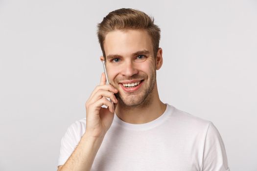Hello im listening. Friendly good-looking smiling blond caucasian guy in white t-shirt answering call, holding smartphone near ear, talking, making order, standing white background.