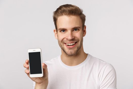 Promo, internet and mobile technology concept. Close-up studio portrait attractive excited and pleased young blond caucasian bearded man in white t-shirt, showing smartphone application screen.