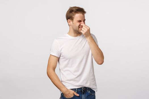 Disappointed and disgusted blond handsome caucasian man close nose with fingers, squinting and turn away from aversion and disgusted, smell something bad, product stinks, standing white background.
