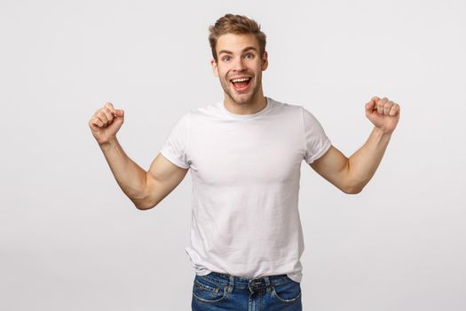 Handsome happy smiling masculine guy, raising hands up fist pump and grinning, stare camera joyful surprise, celebrating team scored, he won big money in lottery, bid, standing white background.