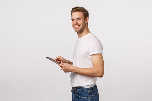 Attractive blond male office assistant, manager, using digital tablet to input or record information, using corporate application, standing profile, turn camera and smiling, white background.