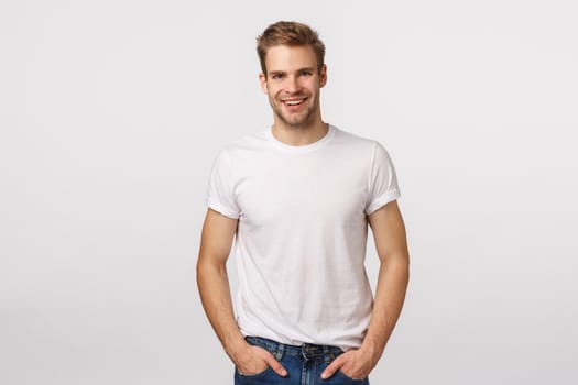 Charming, mature blond male model with bristle, wear white t-shirt, hands in pockets, smiling amused, look entertained, happy expression, agree with friend, standing enthusiastic white background.