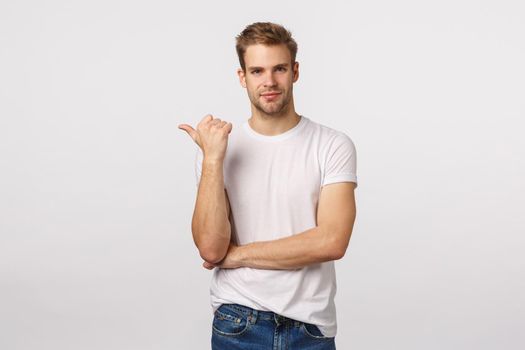 Assertive charming macho man with blond haircut, bristle, white t-shirt, pointing left with thumb and smiling pleased, know exactly what you need, recommend place, company or product, advertising.