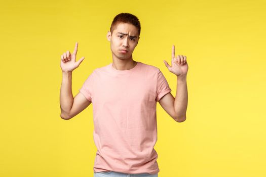 Portrait of unsatisfied, displeased asian guy in pink t-shirt, pointing fingers up, showing disappointed upsetting news, sighing gloomy and look camera upset, stand yellow background.