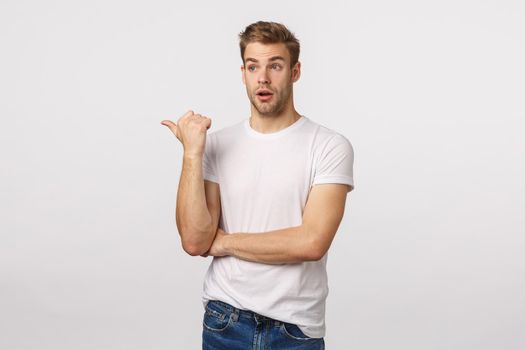 Astounded and impressed cute bearded blond caucasian male in t-shirt, pointing and looking left speechless, getting interested, attend good lecture, standing amazed white background.