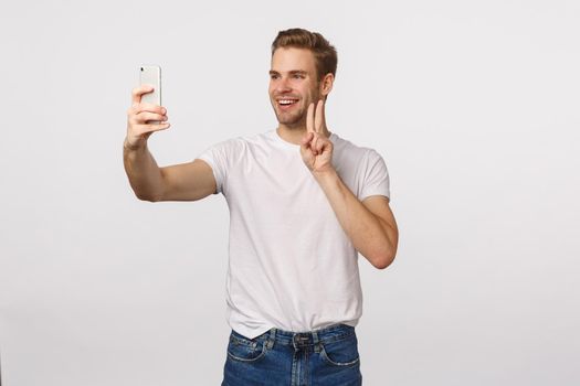 Cheerful man download new app, trying out cute filters as taking selfie on smartphone, making peace sign and smiling joyfully, photographing himself, standing white background.