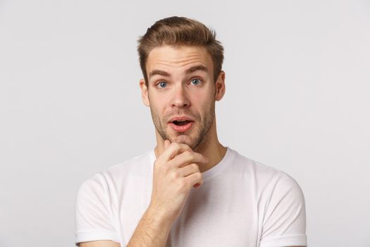 Impressed and surprised, curious good-looking blond bearded male in white t-shirt, touchign chin, open mouth and stare astonished, listen interesting suggestion, stand white background.