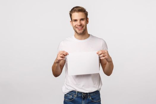 Attractive blond bearded handsome man in white t-shirt, holding blank empty paper as promoting corporate banner, promo, website or advertisement, smiling pleased, white background.