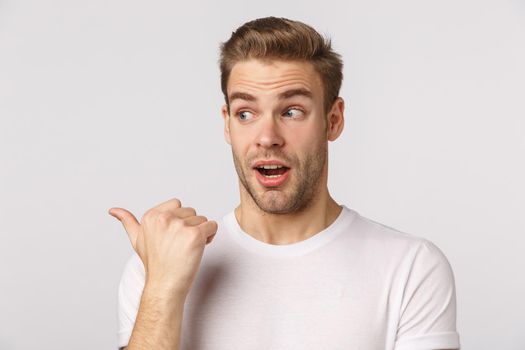 Curious and interested good-looking blond bearded man in white t-shirt, pointing and looking left amazed, see interesting product, promo, asking question, looking intrigued, white background.