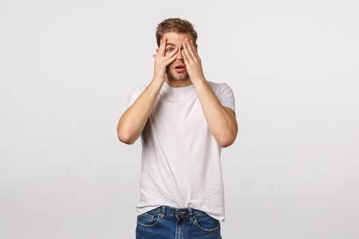 Scared timid and insecure cute blond gay man peeking through fingers with interest and fear, gasping, open mouth, watching scary movie but want see interesting moment, white background.