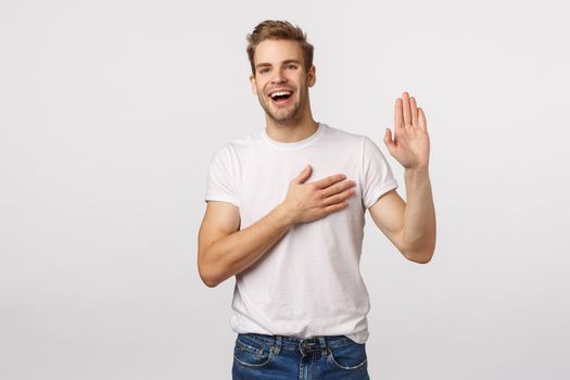 Man giving oath, swear telling truth or be honest. Happy confident and charismatic blond handsome man in white t-shirt, raise one hand and hold another on heart as making promise, white background.