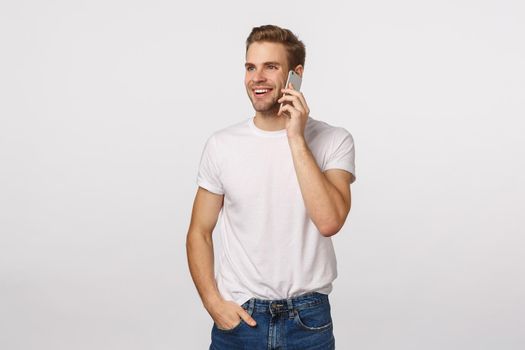 Man calling friend asking girl out. Attractive blond guy in white t-shirt, having conversation, talking on phone, holding smartphone near ear, smiling and look away carefree, white background.