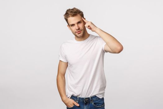 Handsome cheeky young blond guy with bristle in white t-shirt, tilt head and smiling self-assured, assertive, make confident look camera, touching temple, indicate mind, brain, white background.