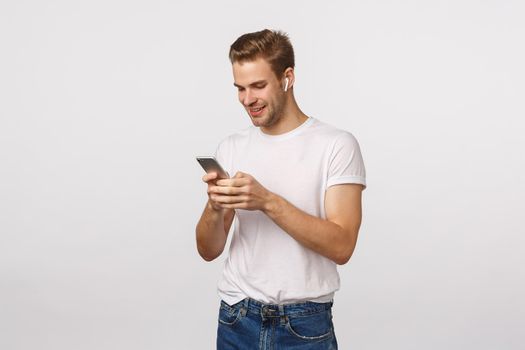 Smiling happy blond european guy in white t-shirt, jeans, holding smartphone, wear wireless headphones, look mobile display, reading message, pick song, white background.