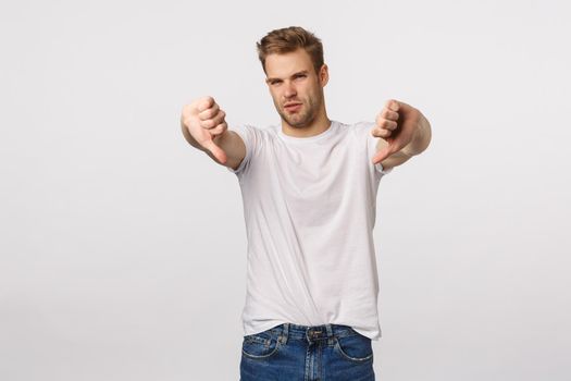 This movie sucks. Displeased and disappointed handsome blond guy give negative feedback, squinting from averstion, shake head in no, never, make thumbs-down gesture, standing white background.