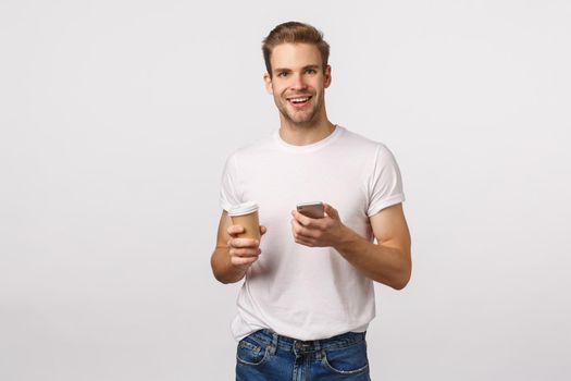 Cheerful good-looking modern blond man with bristle, holding paper cup of coffee, drinking tea and holding smartphone, messaging, smiling camera, browsing internet, shopping online during break.