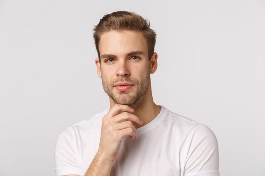 Hmm let me think. Pensive good-looking blond young bearded man in white t-shirt, touching chin and squinting intrigued, thinking, pondering what choice make, standing thoughtful.