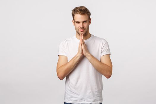 Determined, confident lucky good-looking sassy blond caucasian man, white t-shirt, press hands in pray, hope gesture, look camera self-assured and smiling, relish good deal, white background.