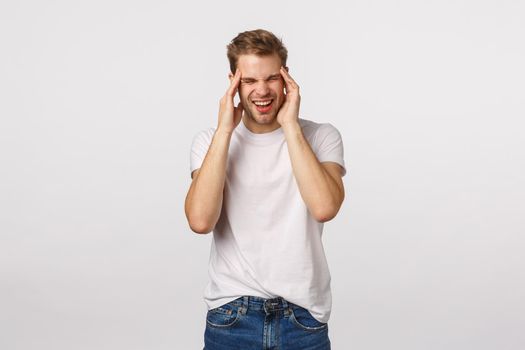 Man suffering headache after hangover, yesterdays party. Handsome blond man complain on migraine, squinting from pain, grimacing bothered, touch temples, have painful feeling, white background.