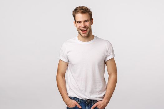 Amused and flirty, coquettish macho man with blond haircut, white t-shirt, bristle, smiling and staring with affection or temptation, like what he see, feeling sexy and confident, white background.