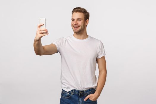 Say cheese. Friendly handsome smiling, stylish blond modern guy with bristle, extend hand holding smartphone, taking selfie and smiling, stream vlog online using mobile phone, white background.