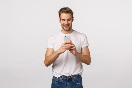 Enthusiastic handsome and cute blond boyfriend taking photo for girlfriend, shooting event, record video on smartphone, holding mobile phone and looking at screen with pleased smile, white background.