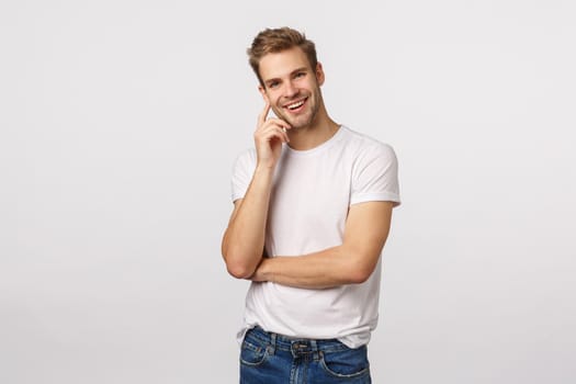 Charming handsome smiling blond caucasian man grinning at camera, touching cheek and tilt head as listening interesting story, agree with friend, like what hear, standing pleased white background.