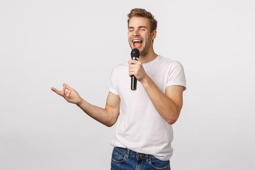 Carefree and joyful good-looking bearded blond guy in white t-shirt, holding microphone singing karaoke, gesturing, close eyes and smiling as feeling happy enjoy, standing white background.