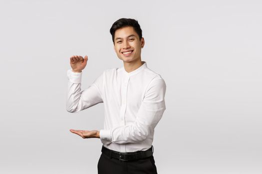 Joyful good-looking asian male entrepeneur, employee in white shirt, pants, showing big box with pleased expression, holding product or something large and satisfying, stand white background.