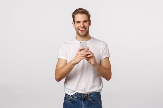 Excited, happy and cheerful blond stylish handsome man in white t-shirt, record concert on phone, taking picture, photoraphing, making good shot with smartphone camera, smiling thrilled.