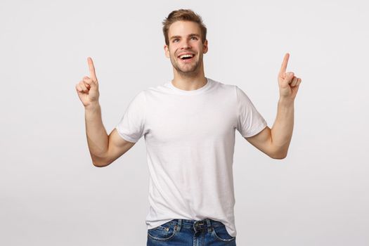 Impressed, carefree delighted, smiling young man feeling joy or happiness, grinning astounded or wondered, pointing looking dreamy up, watching amusing scene, standing white background charmed.