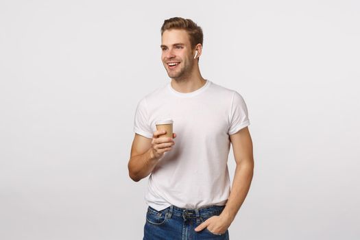 Modern life, lifestyle and people concept. Attractive blond caucasian guy holding take-away paper cup, enjoy coffee and talking phone via wireless headphones, smiling look away, white background.