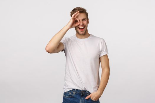Happy cheerful blond male model with bristle, wear white t-shirt, looking through circle or okay sign with satisfied smile, leave positive feedback, give approval, like product, white background.