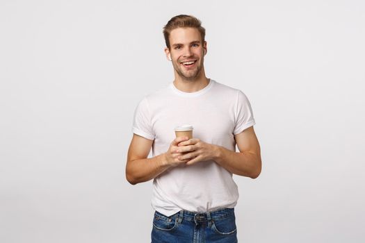 Lifestyle, technology and urban concept. Handsome smiling blond guy holding paper cup of coffee, order take-away, listen music in wireless earphones, use headphones to talk friend via hands busy.