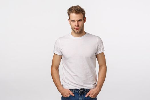 Angry, judgemental handsome macho blond male model in white t-shirt, look offended or angry, squinting aggressive, judging or being disappointed, standing suspicious, have doubts, white background.