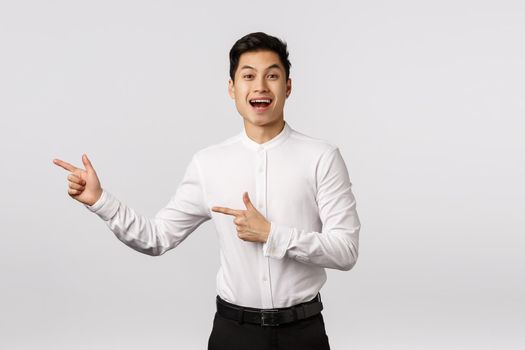 Impressed, happy smiling asian elegant guy, male student graduating, wear formal outfit to after-party, business meeting, pointing left, grinning joyfully, recommend visit internet page.