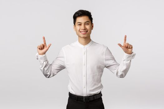 Hansdome asian businessman in white shirt, black pants, pointing fingers up and smiling with pleased, satisfied or glad expression, showing product, promoting corporate banner, white background.