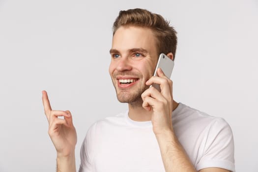 Close-up blond handsome man with blue eyes, white t-shirt, holding smartphone near ear, gesturing finger, smiling pleased, make order, confirm online shopping, talking with friend, white background.