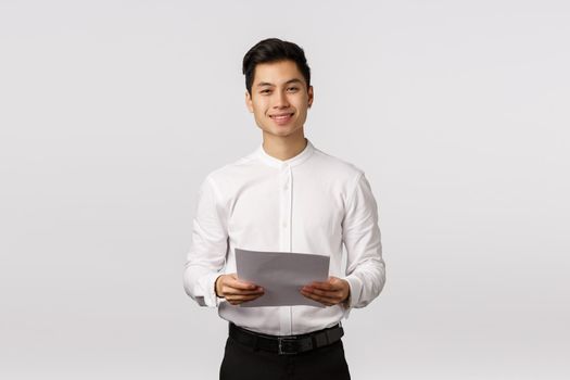 Handsome elegant asian young male coworker, businessman in white collar shirt, black pants, holding papers and smiling camera, successful entrepreneur prepare report, reading speech.