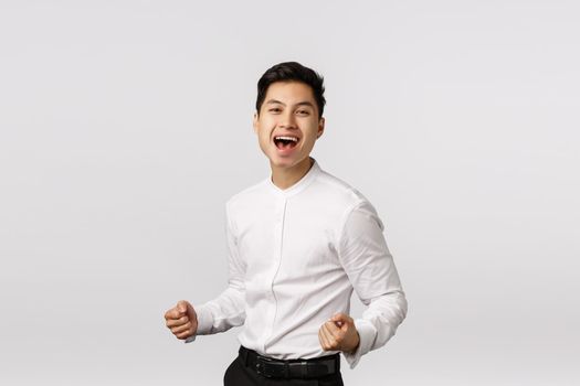 Delighted, successful young asian businessman clench fists, pump it up and smiling pleased, say yes, celebrating win, placed good bid, achieve goal, rejoicing receive prize, standing white background.