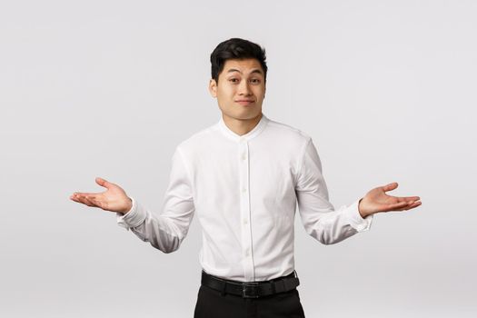 Cant help you sorry. Handsome asian businessman shrugging, spread hands sideways dismay and wonder, frowning and smirk awkward, dont know anything, clueless, unable to answer, white background.