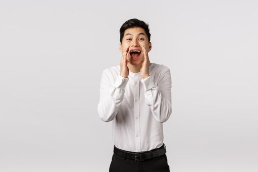 Hey you come here. Cheerful smiling happy asian guy searching new employees to company, hire, holding hands near opened mouth shouting name, searching someone, stand white background.