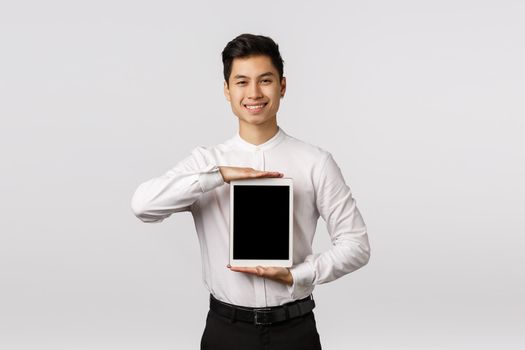 Satisfied handsome asian businessman, showing digital tablet screen over chest and smiling pleased, give promo, advertise gadget application or shopping site, showing corporate link.
