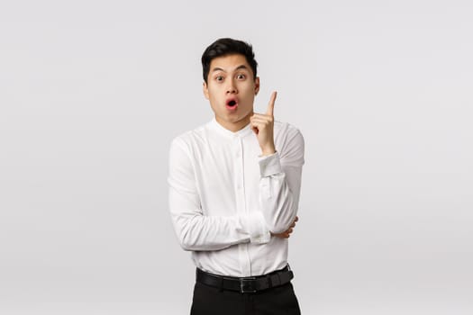 Excited, fascinated asian businessman found solution, got excellent idea, raise index finger in eureka, got it gesture, open mouth, say his suggestion, standing white background thrilled.