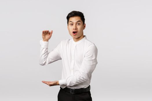 Impressed lucky handsome asian man in white shirt, pants, open mouth say wow astonished, showing big box, large product with hands gesture, standing fascinated white background.