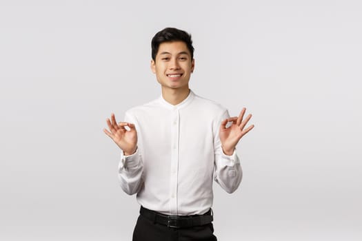 Success, achievement and gestures concept. Handsome young male asian entrepreneur showing good, alright or okay gesture, smiling in approval, like idea, pleased with results, white background.