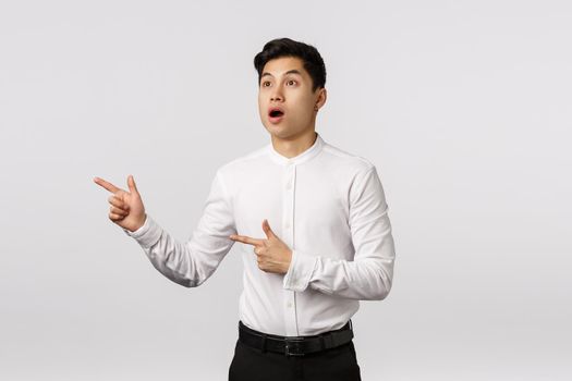 Speechless, surprised and impressed attractive asian young businessman in white shirt, black pants, open mouth fascinated, man attention captured on awesome promo, look pointing left.