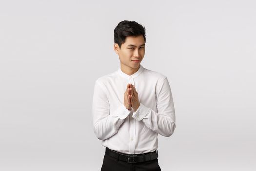 Mysterious and devious young good-looking asian businessman in white shirt, pants, rub hands and squinting sly, have secret evil plan, relish good deal, make money, standing white background.