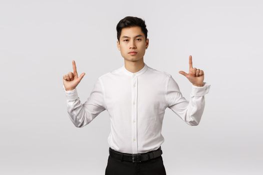 Serious-looking professional, successful asian young male entrepreneur pointing up and looking camera assertive, know exactly what customer need, suggest product, give perfect variant.